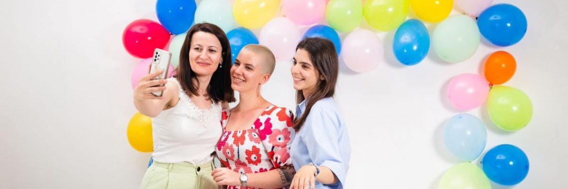Three women smiling and taking a selfie against a vibrant balloon backdrop.